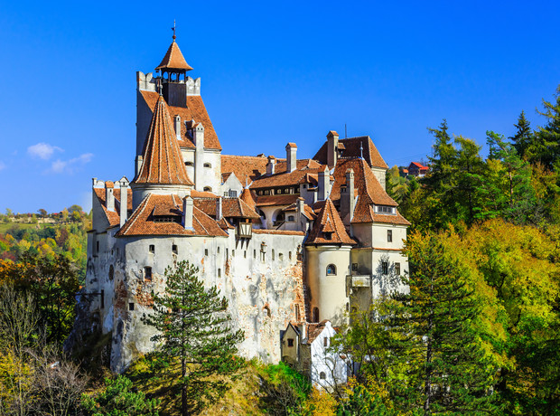 A large, historic castle with red-tiled roofs and towers sits atop a forested hill, surrounded by colorful autumn trees under a clear blue sky.